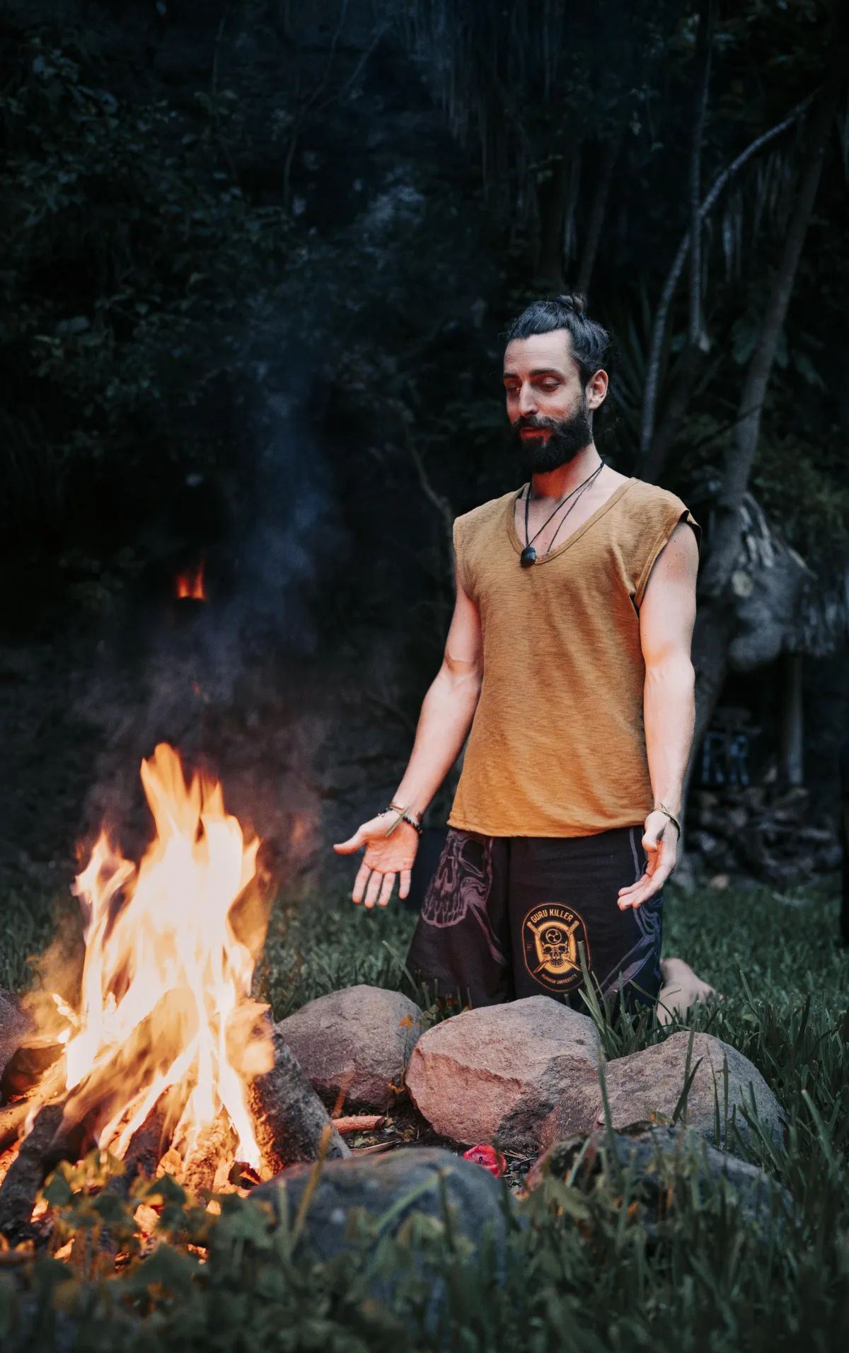 A man kneeling beside a fire in the jungle at dusk, palms open to the flames