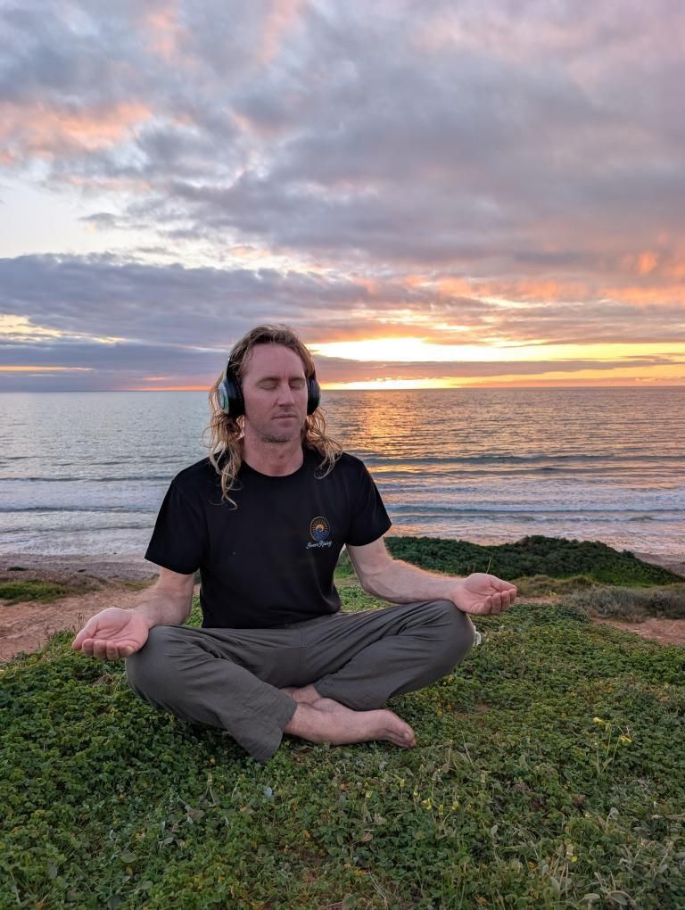 Joel Ley sitting cross-legged on a clifftop at sunset above the ocean, eyes closed, wearing headphones