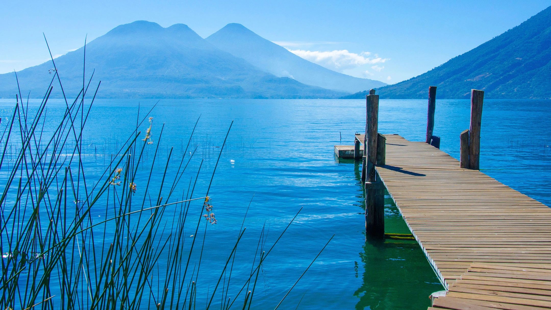 A wooden jetty reaching into the blue water of Lake Atitlán with volcanoes behind