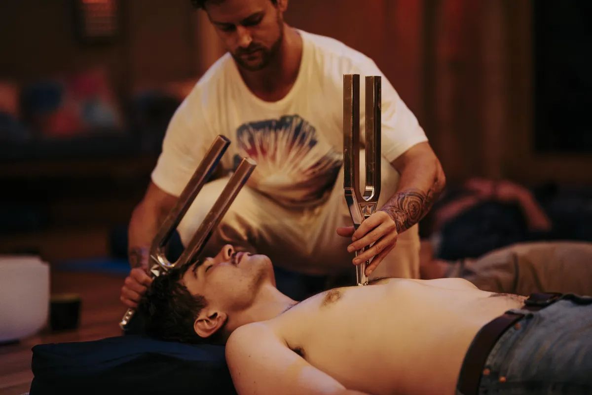 A facilitator holding two tuning forks above a person resting on a mat in a softly lit room